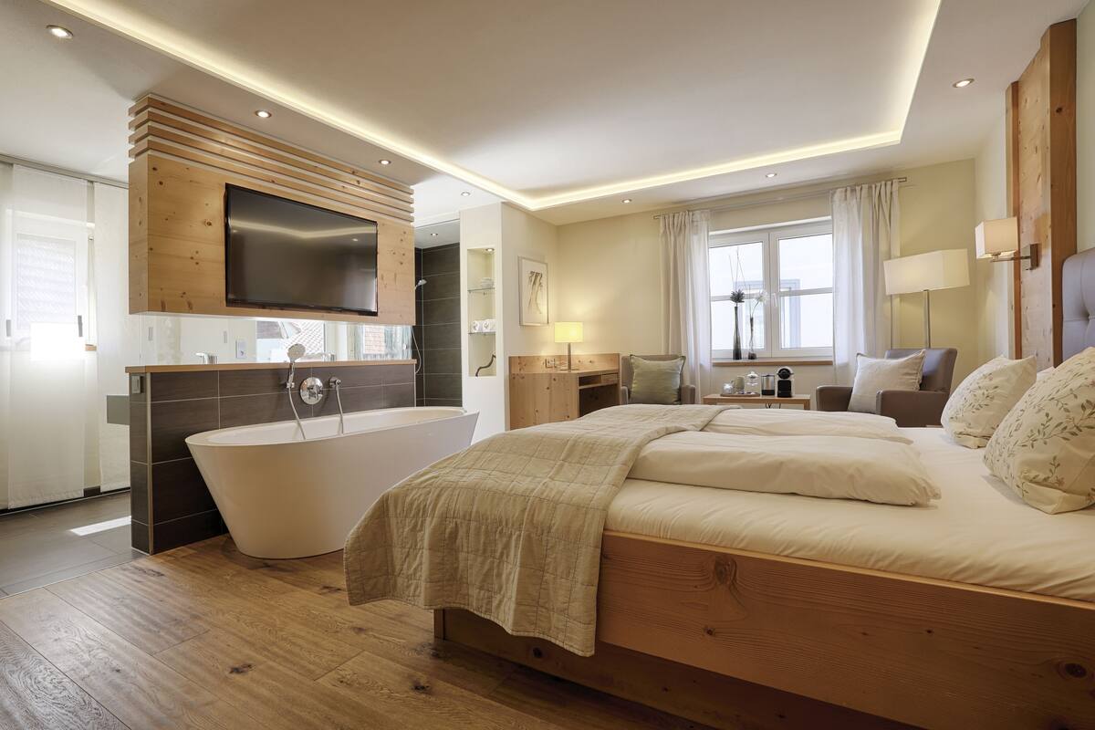 Quarto moderno com cama grande, banheira independente e TV de tela plana fixada em um painel de madeira.