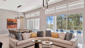 A spacious living room with a large L-shaped sofa, a dining area with a table and chairs, and large windows offering a view of a swimming pool and greenery. - Grand Home w Karaoke RM, Piggy Playhouse BR, Theater, Arcade & Gym, Near Disney (Davenport)