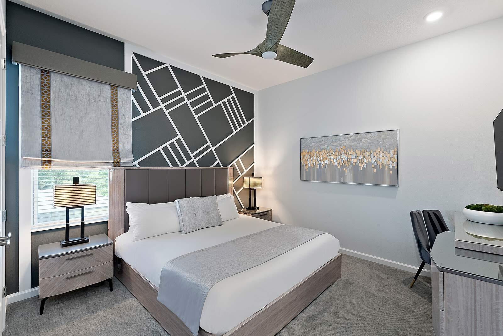 A modern bedroom with a large bed, a desk, a chair, a ceiling fan, and a wall with geometric patterns.