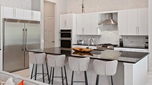A modern kitchen with white cabinetry, a stainless steel refrigerator, a built-in oven, a gas stove, and a central island with bar stools. - Grand Home w Karaoke RM, Piggy Playhouse BR, Theater, Arcade & Gym, Near Disney (Davenport)