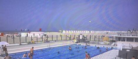 A swimming pool with multiple lanes, surrounded by a fence, and a beach area in the background.