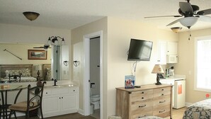 A bedroom with a bed, a desk, a chair, a TV, a ceiling fan, and a bathroom with a toilet and sink.