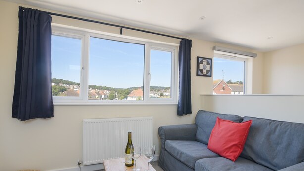 A living room with a grey sofa, a small wooden table with a wine bottle and glasses, a radiator, and a view of houses through large windows. - Ayla (Lyme Regis)