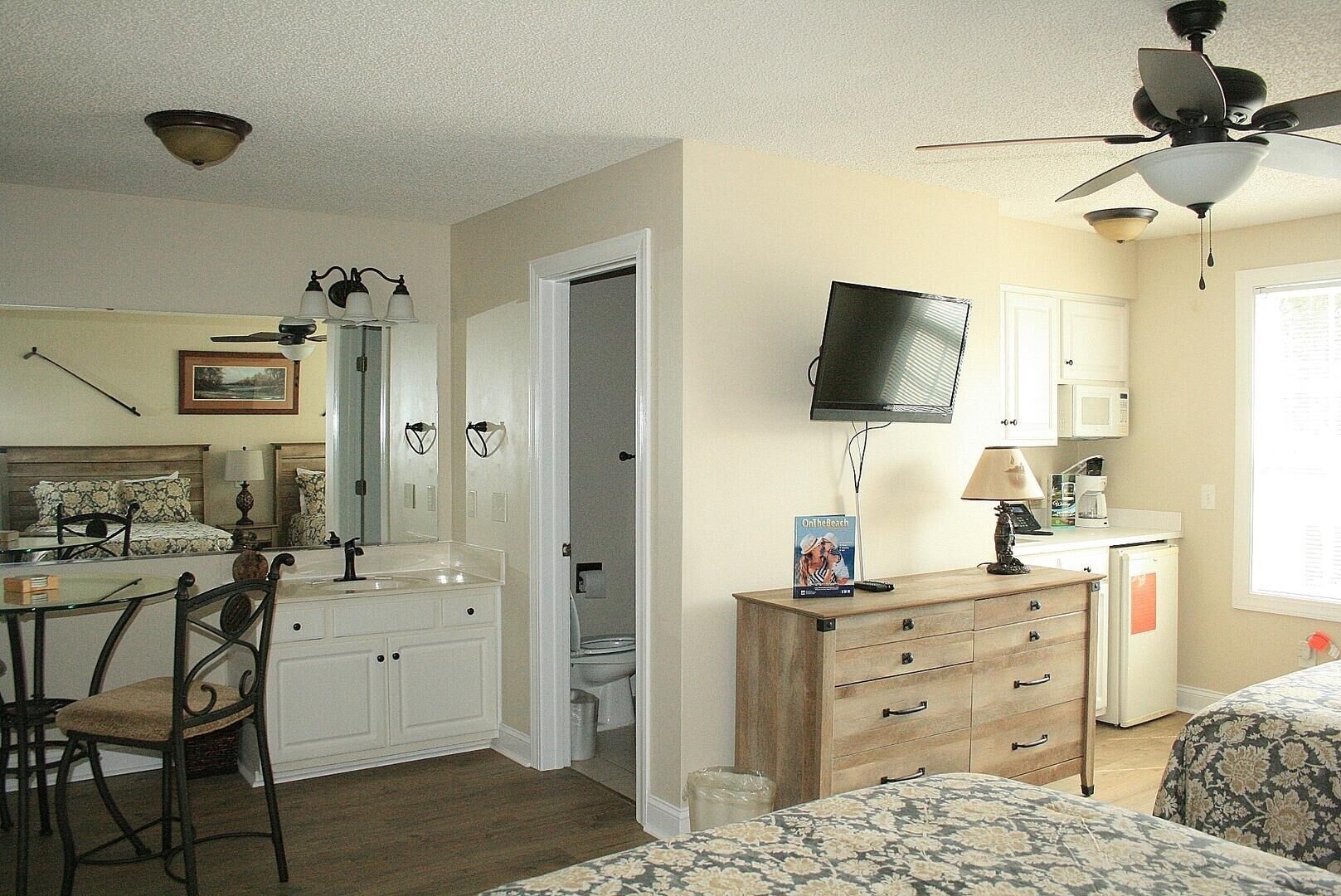 A bedroom with a bed, a desk, a chair, a TV, a ceiling fan, and a bathroom with a toilet and sink.