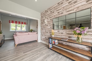 Uma sala de estar moderna com parede de pedra, uma janela grande com cortina em vermelho e branco, sofá rosa, estante de madeira e um vaso com flores.