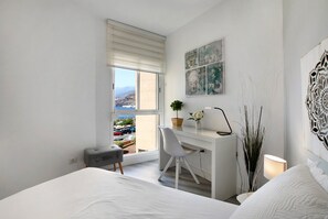 Une chambre à coucher comprenant un lit, un bureau, une chaise, une fenêtre donnant sur un beau paysage et un mur décoré d’œuvres d’art.