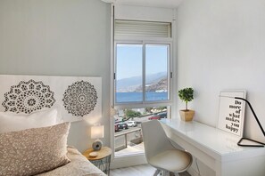 A bedroom with a large window, a bed, a desk, a chair, and decorative wall art.