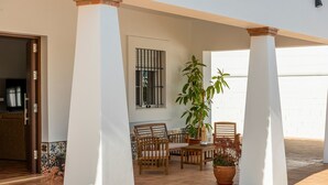A covered outdoor seating area with wooden chairs and tables, a potted plant, and a window with bars. - Villa "Casa Brisa del Valle" with Private Pool, Covered Terrace & Wi-Fi (Sevilla)
