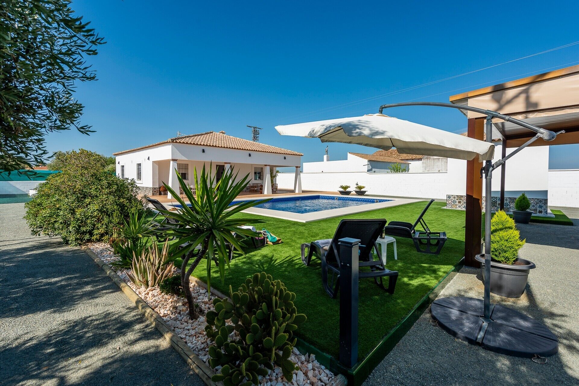 Zona de piscina con una casita, mobiliario exterior y un espacio de estar techado.