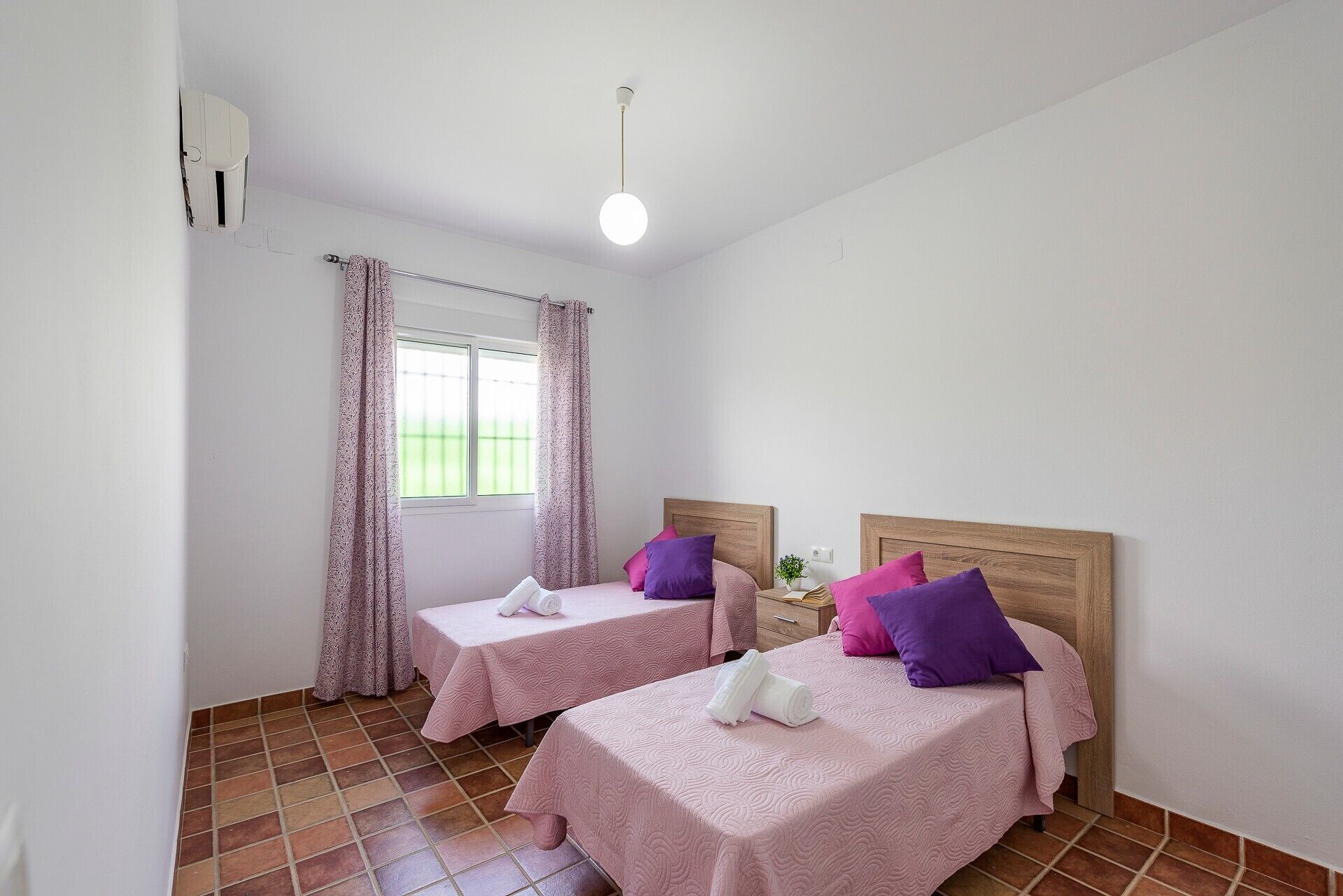 A room with two beds, a window with curtains, a ceiling light, and a tiled floor.