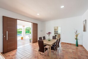 Ein Essbereich mit Holztisch und -stühlen, Blick auf einen Garten durch eine offene Tür, und eine Küche, die durch eine weitere offene Tür sichtbar ist.