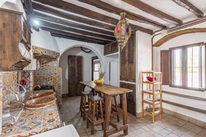 A rustic kitchen with a wooden table, stools, a hanging light fixture, and a tiled backsplash.