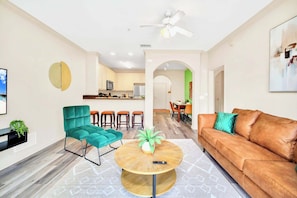Una sala de estar moderna con un sofá, un sillón color turquesa, una mesita de centro y una cocina al fondo.