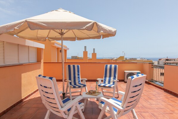 Eine Terrasse mit zwei gestreiften Stühlen, einem Tisch mit einer Flasche und Snacks sowie einem großen Sonnenschirm.