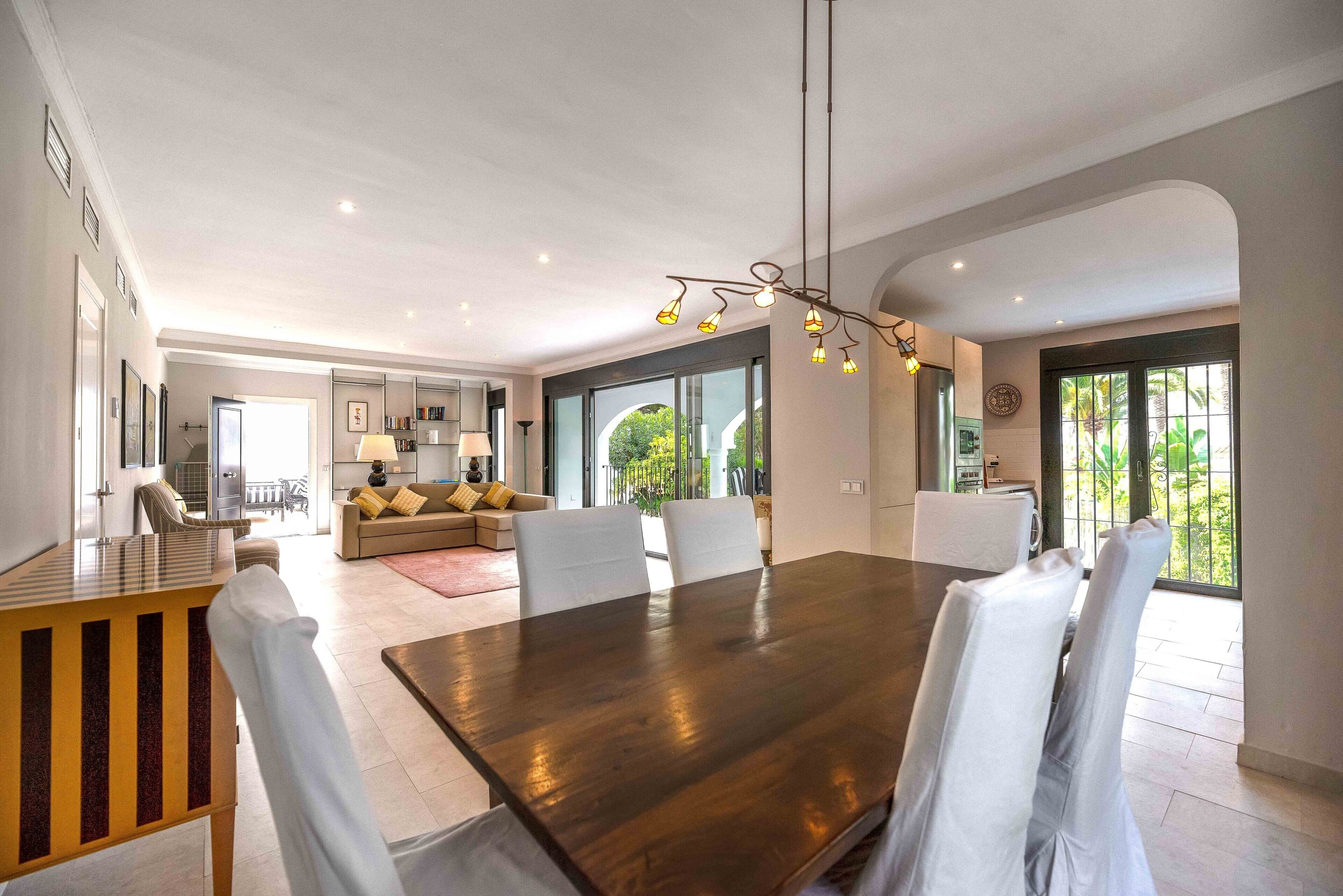 Un vaste espace dédié à la salle à manger, avec une grande table en bois et des chaises blanches, une cuisine moderne en arrière-plan et de grandes fenêtres offrant une vue sur la verdure.