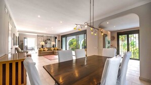 Un vaste espace dédié à la salle à manger, avec une grande table en bois et des chaises blanches, une cuisine moderne en arrière-plan et de grandes fenêtres offrant une vue sur la verdure.