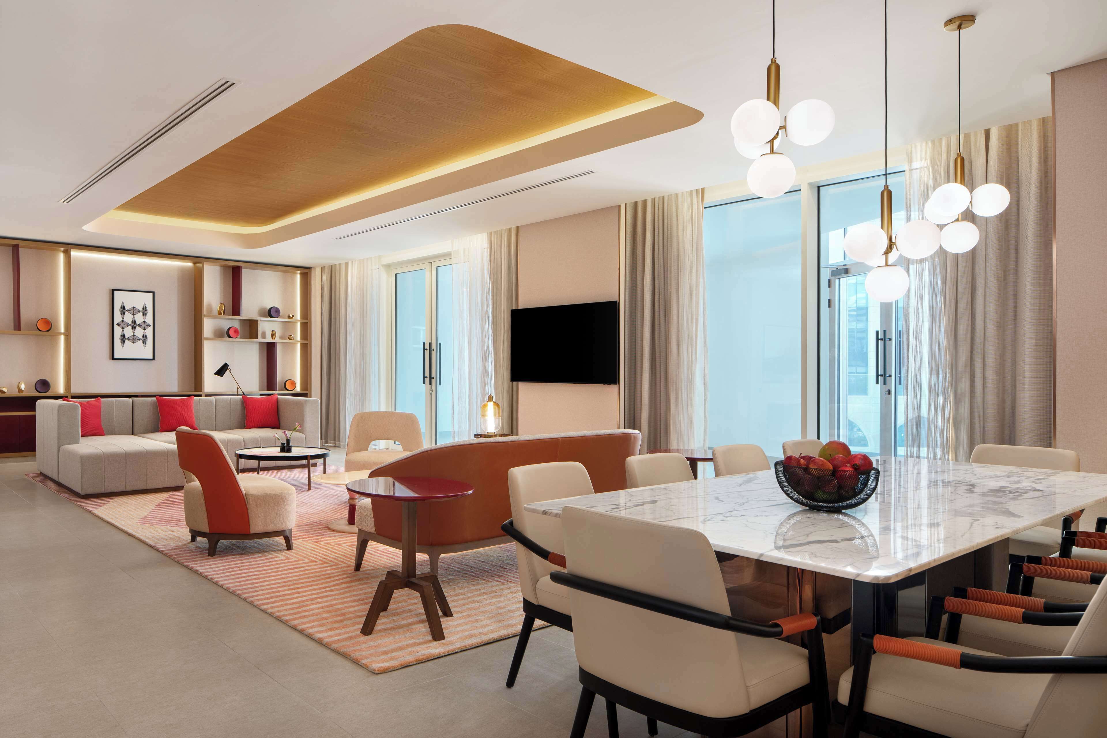 Habitación de hotel moderna con una amplia mesa de comedor, asientos cómodos, un televisor de pantalla plana y un ventanal con cortinas.