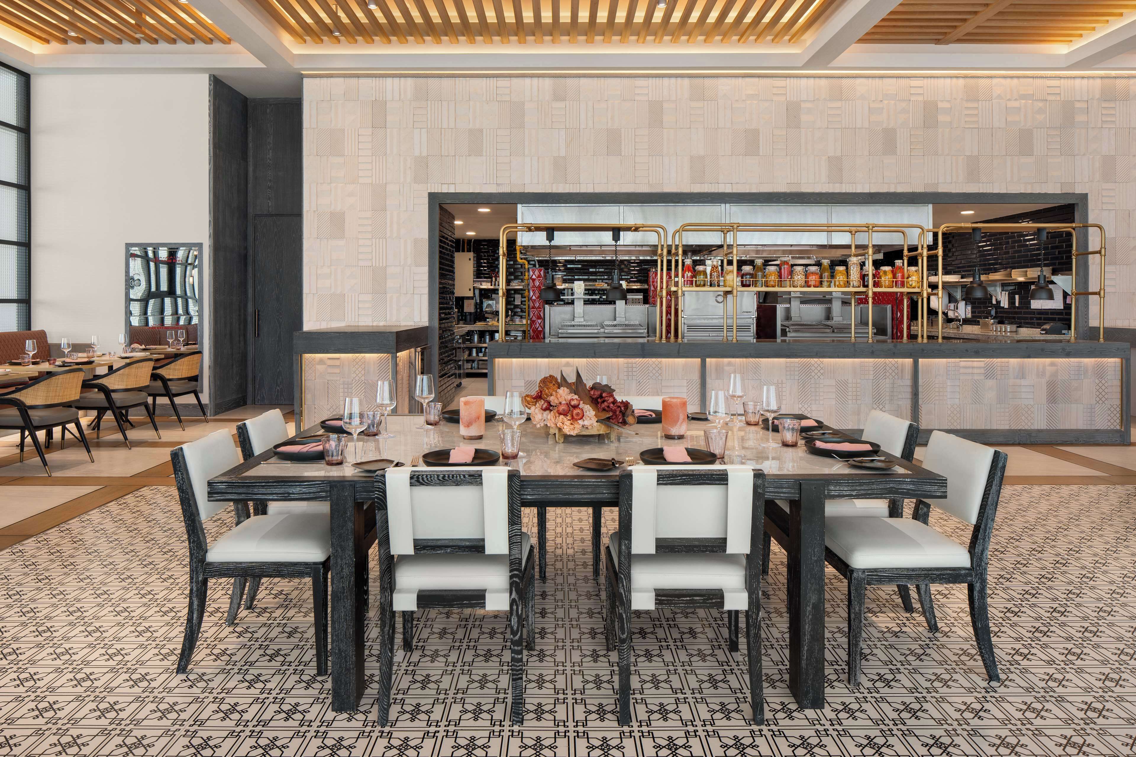 A dining area with a long table set for six, surrounded by chairs, in a modern interior with a patterned floor and a kitchen visible in the background.