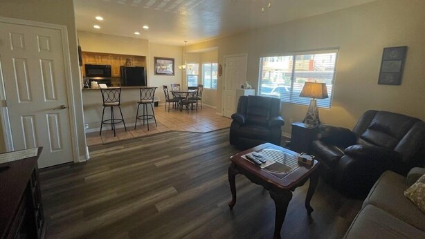 A spacious living room with a dark wood floor, a ceiling fan, a dark leather sofa, and a coffee table. There is a kitchen area with a microwave and a refrigerator in the background. - Kings Court by Mesquite Vacation Rentals with Community Pool and $510 in free daily activities (Mesquite)