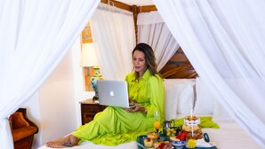 Una persona con un vestido verde brillante sentada en una cama con una computadora portátil, rodeada por una bandeja de desayuno con diversos alimentos y un oso de peluche.