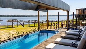 Zona de piscina con vistas a un lago, terraza de madera y sillones de descanso.