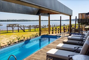 Zona de piscina con vistas a un lago, terraza de madera y sillones de descanso.
