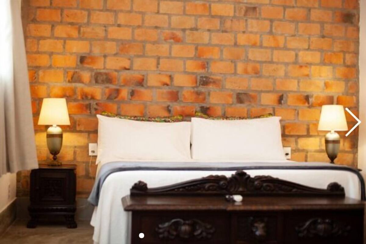 A hotel room with a brick wall, a bed with white bedding, two bedside tables with lamps, and a dark wooden dresser.