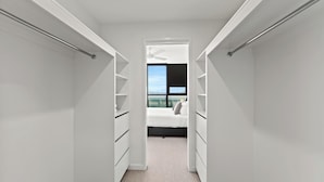 Ein schmaler Durchgangskasten mit weißen Regalen und Schubladen, der in ein Schlafzimmer mit Bett und Fenster mit Blick auf eine Stadtlandschaft führt.