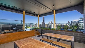 An outdoor seating area with wooden benches and tables, a glass railing, and a city view. - Fortitude Valley Apartments By Cllix (Brisbane)