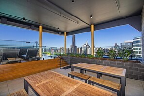 Un espace extérieur aménagé avec des bancs et des tables en bois, une rambarde en verre et une vue sur la ville.