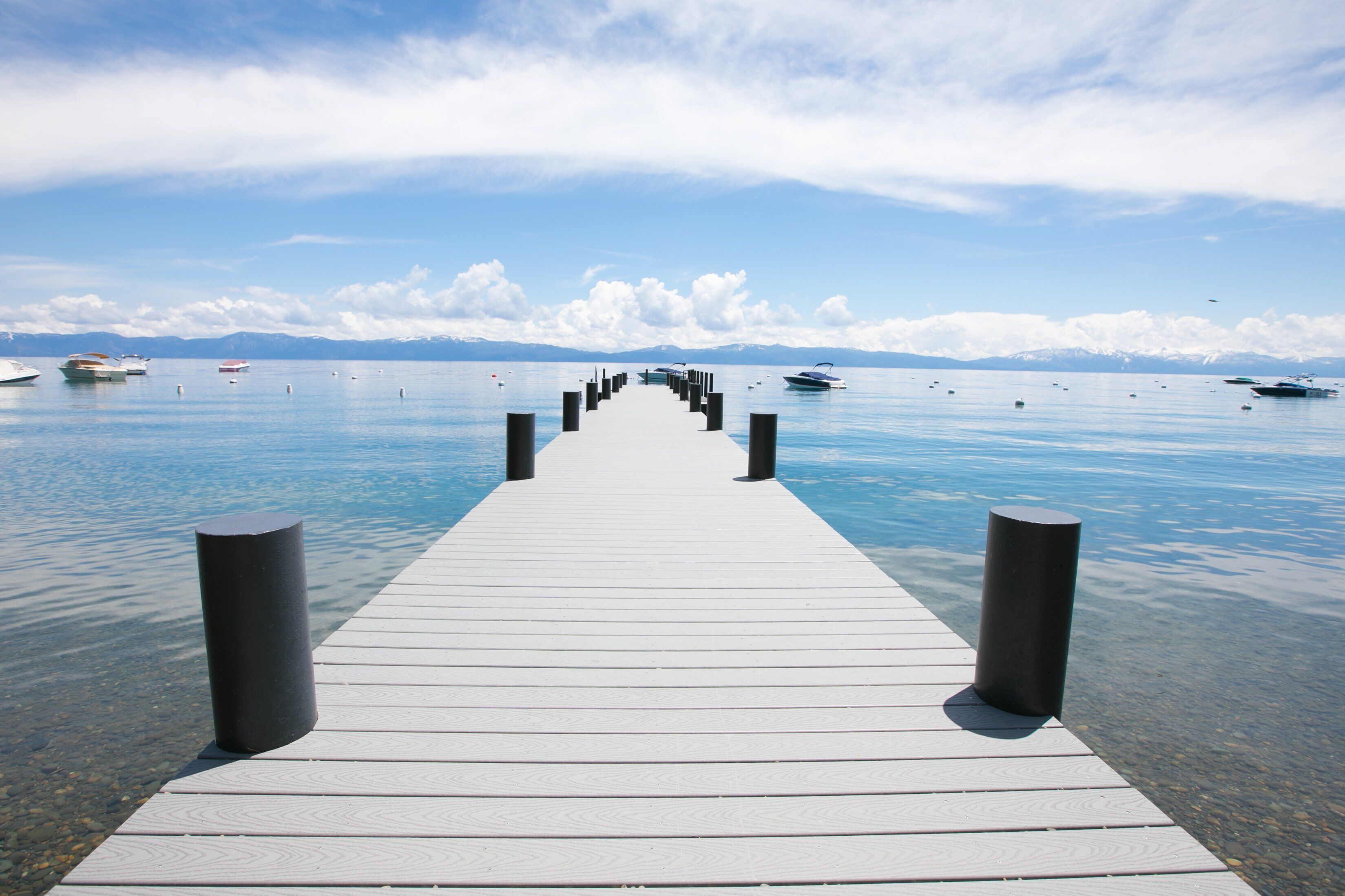 Un muelle de madera que se adentra en aguas tranquilas, con barcos y montañas al fondo.