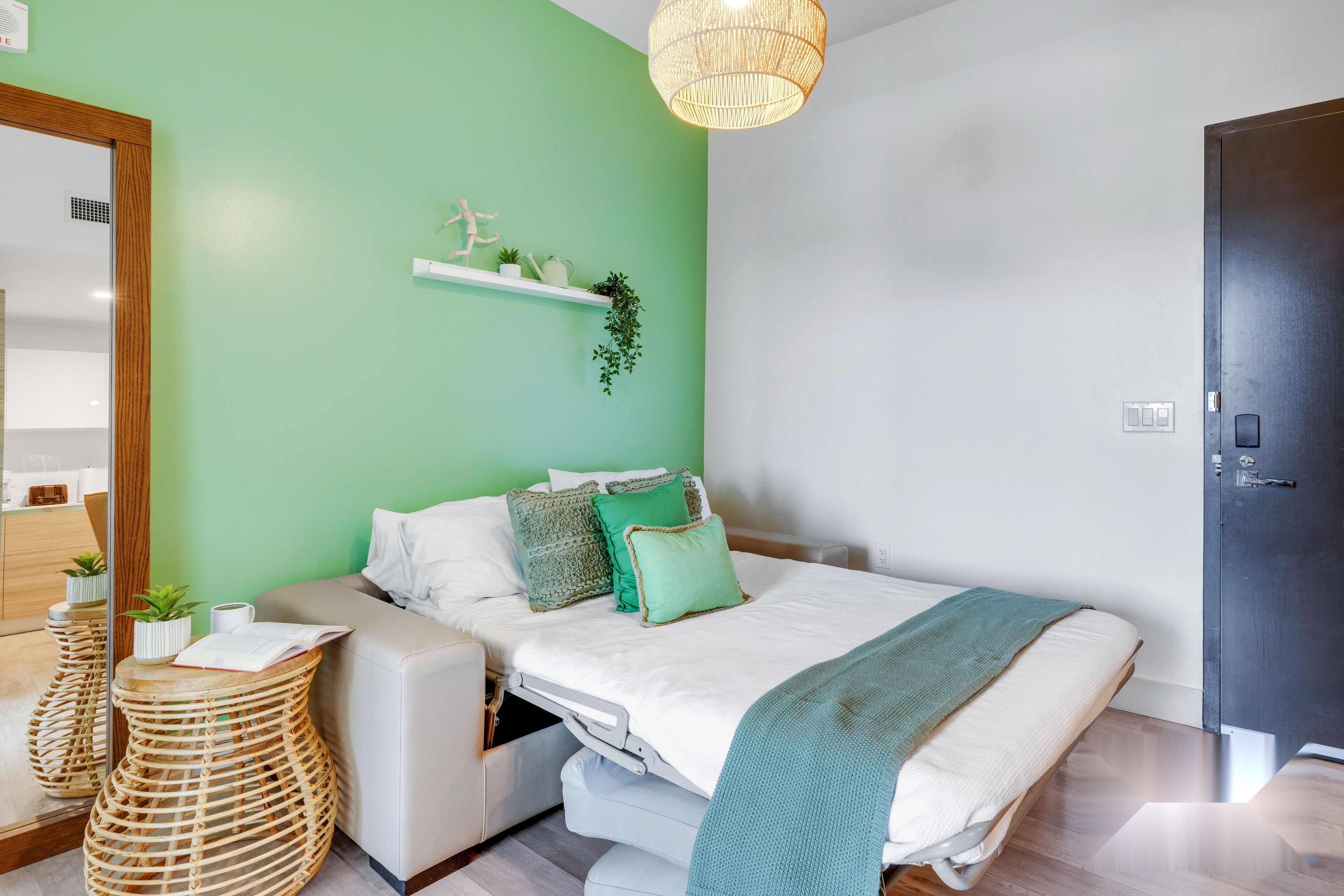 Un dormitorio moderno con una pared de acento verde, una cama con ropa de cama blanca, una mesita de noche con una planta y una lámpara colgante.
