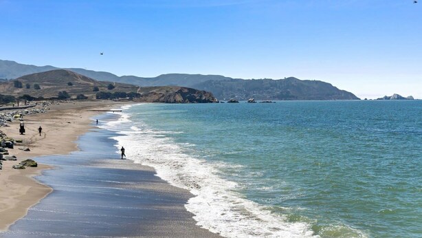 A beach with clear blue water, rocky shoreline, and distant hills.