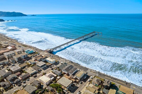 A coastal town with a pier extending into the ocean, residential buildings, and a clear blue sky.