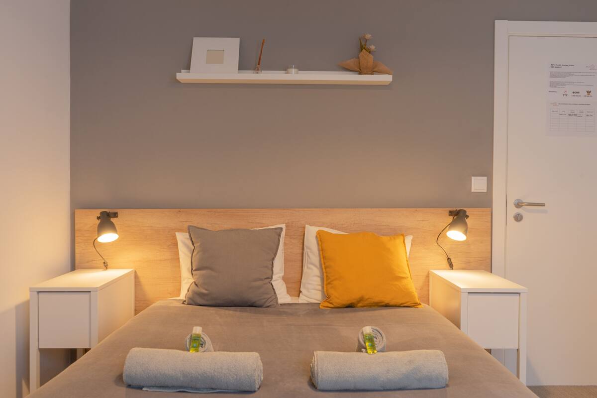 A neatly made bed with two pillows, a bedside table with a lamp, and a shelf above the bed.