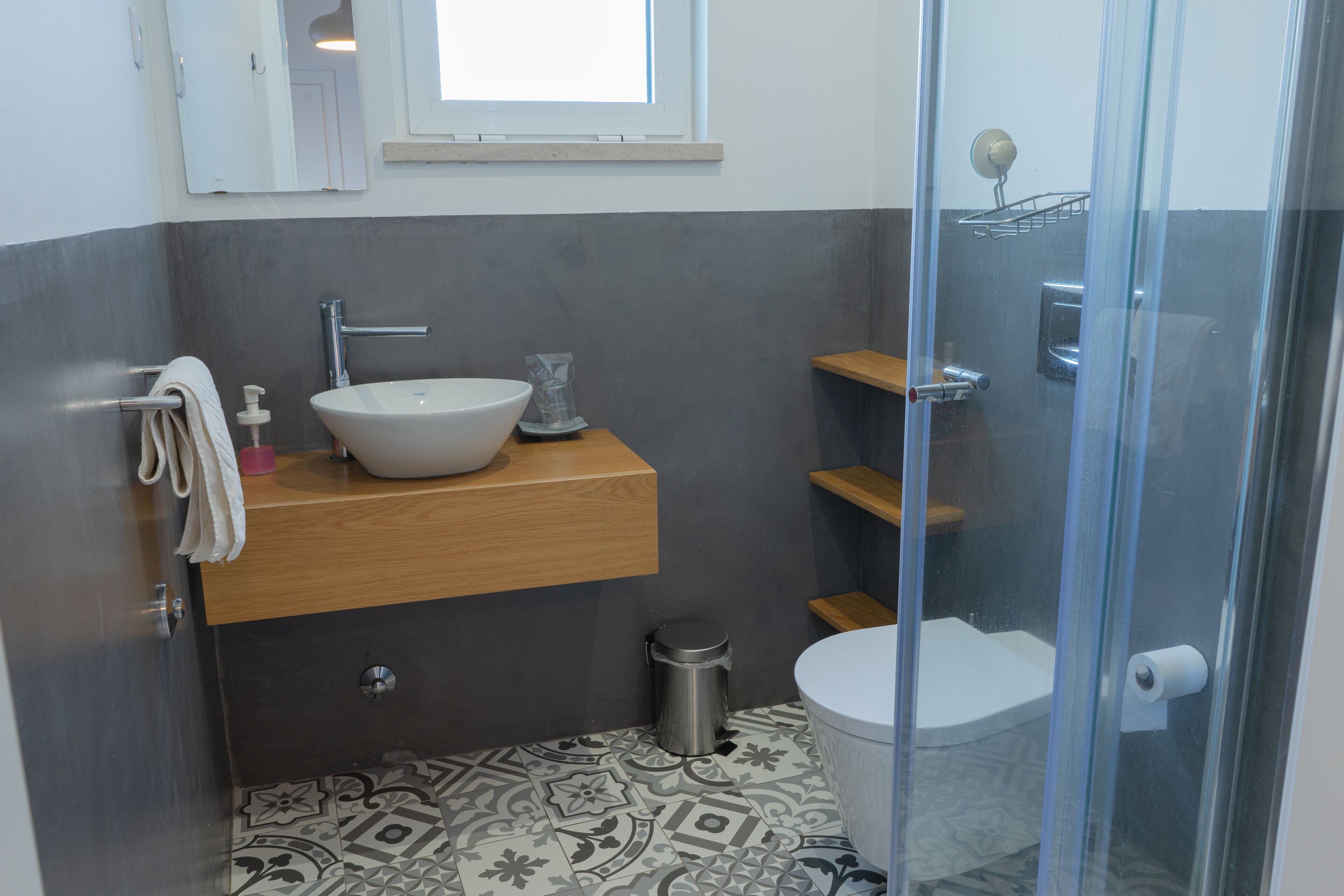 Un baño moderno con lavamanos blanco, tocador de madera, baldosas de suelo estampadas, mampara de cristal para la ducha y un inodoro.