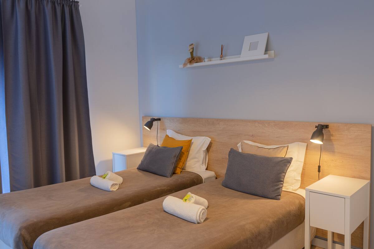 A hotel room with two beds, a nightstand with a lamp, a shelf with decorative items, and a curtain.
