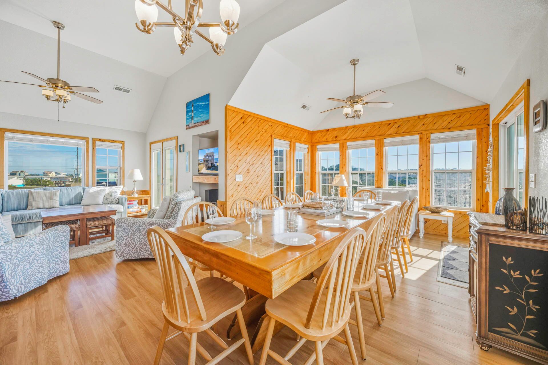 A spacious dining area with a long wooden table set for six, surrounded by wooden chairs. There are two ceiling fans, a large window with a view, and a wooden wall paneling.