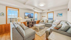 A spacious living room with two sofas, a coffee table, and a bookshelf. Large windows offer views of the outdoors. - Soundfront Home with Private Pool, Hot Tub, and Private Windsurfing Access (Waves)