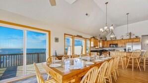 A spacious dining area with a large wooden table set for six, wooden chairs, and a view of the ocean through large sliding glass doors. - Soundfront Home with Private Pool, Hot Tub, and Private Windsurfing Access (Waves)