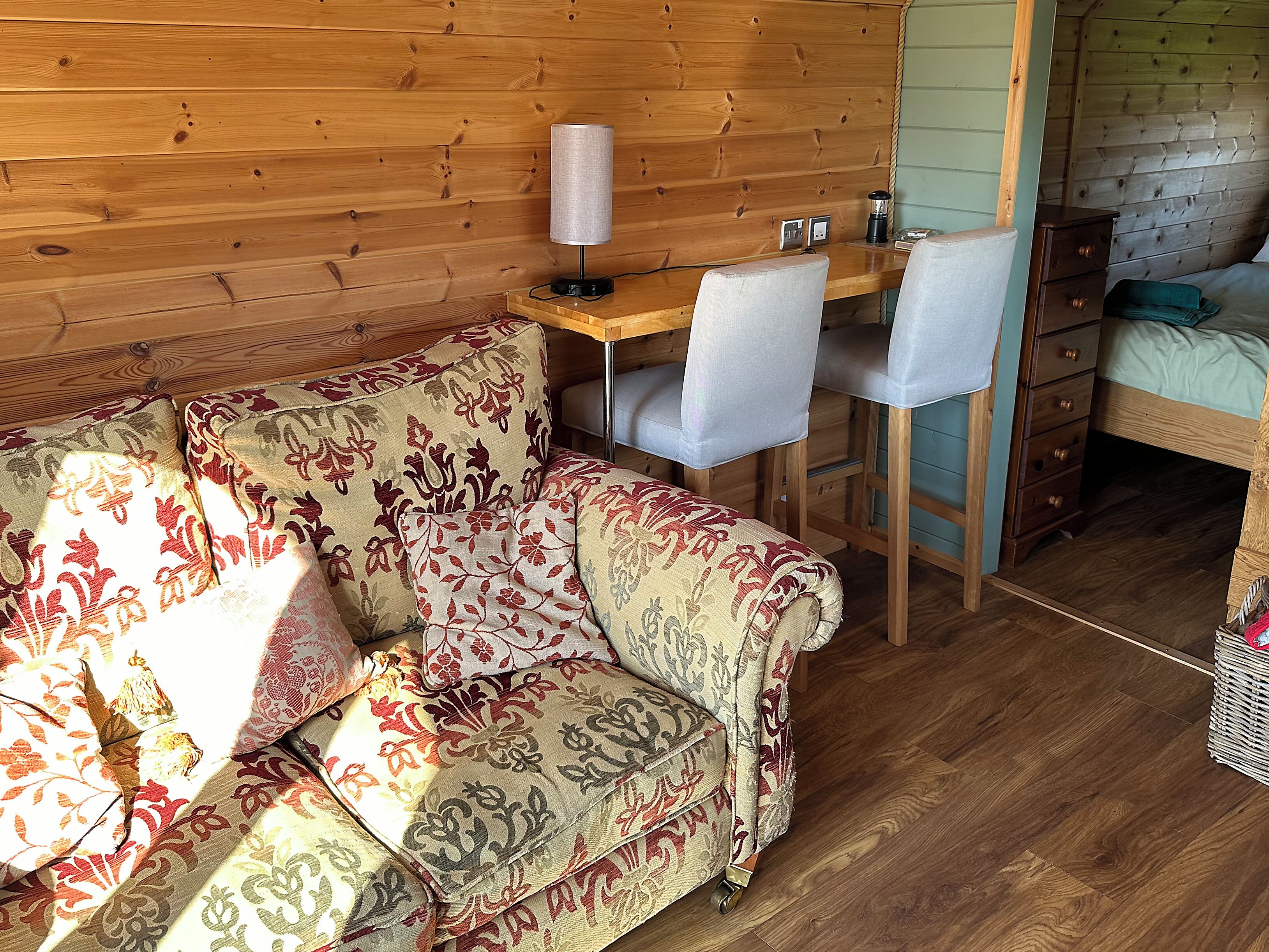 A cozy room with a wooden floor, a patterned sofa, a small desk with a lamp, and a bed with a green blanket.