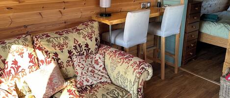 A cozy room with a wooden floor, a patterned sofa, a small desk with a lamp, and a bed with a green blanket.