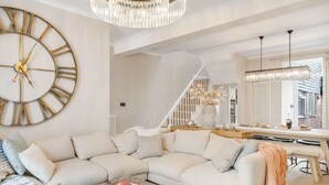 Un salon moderne avec une grande horloge murale, un canapé blanc, une table basse et un coin repas agrémenté d’un lustre.