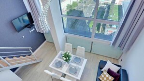 A modern apartment with a dining area, a TV mounted on the wall, and a large window offering a city view.