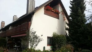 Un edificio blanco con techo inclinado, un balcón de madera con flores y una ventana pequeña.