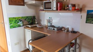 Una cocina compacta con microondas, un refrigerador pequeño y un lavamanos. Hay una mesa de madera y una silla, junto con un estante sobre la encimera con varios objetos.