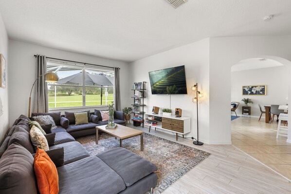 A modern living room with a large sectional sofa, a coffee table, and a TV mounted on the wall. There is a bookshelf with books and decorative items, and a dining area in the background.