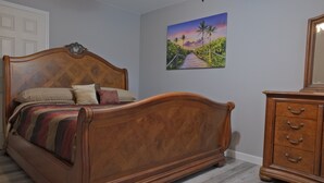 A bedroom with a wooden bed, a dresser, and a painting of a scenic pathway. - Awalk away from Busch Gardens and Adventure Island (Tampa)