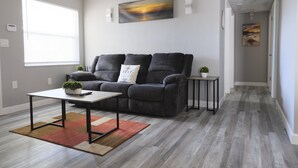 A modern living room with a grey sofa, a coffee table, and a small side table. There is a painting on the wall and a window with blinds. - Awalk away from Busch Gardens and Adventure Island (Tampa)