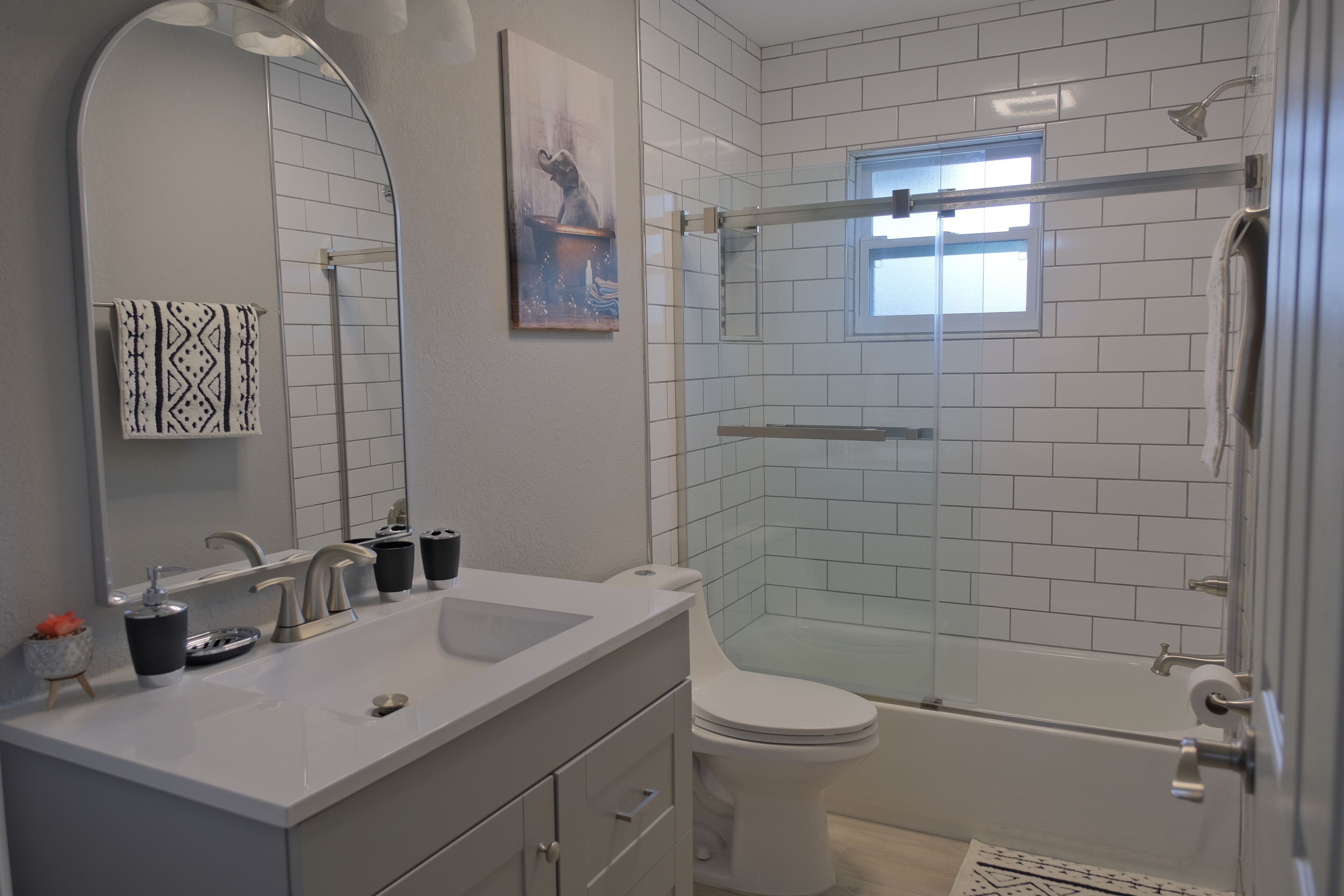 Een moderne badkamer met een witte wastafel, spiegel en toilet. Het heeft een douche met glazen wand en een klein raam.